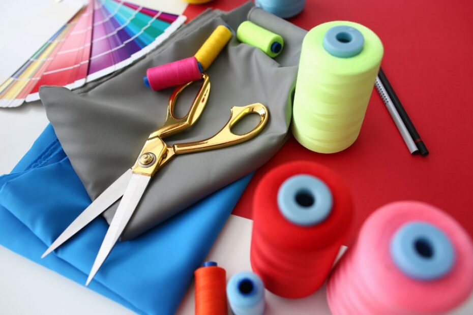 Tesoura dourada sobre tecidos coloridos, carretéis de linha em tons vibrantes e cartela de cores ao fundo, representando materiais para costura e artesanato criativo.