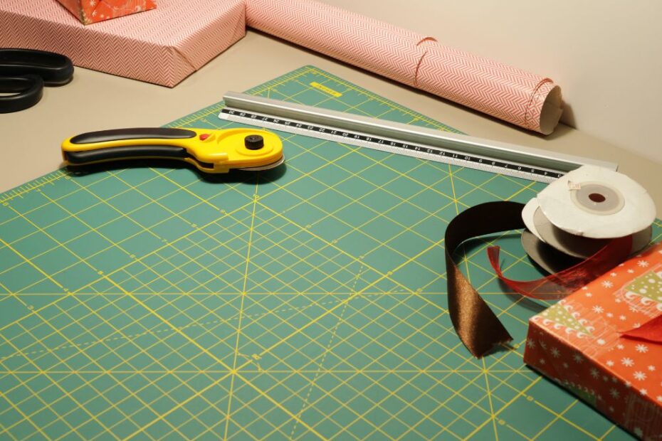 Mesa de trabalho com base de corte verde, cortador circular, régua metálica, fitas decorativas e papel de presente para embalagem e artesanato.